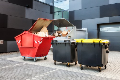 The image shows three large wheelie bins positioned outdoors on a paved area adjacent to a modern building with dark grey exterior panels. The leftmost bin is red with a partially open lid, revealing a stack of flattened cardboard boxes and other paper waste inside. The middle bin is black with a closed yellow lid, and the rightmost bin is also black with a closed black lid. The bins are situated in a neat, urban environment, typical of commercial waste collection sites, with no visible waste scattered around. The red bin's open lid and contents suggest ongoing rubbish collection or disposal, in line with services offered by Commercial Waste Eltham in Eltham. The setting appears clean and organized, highlighting the importance of proper waste management solutions in the area.
